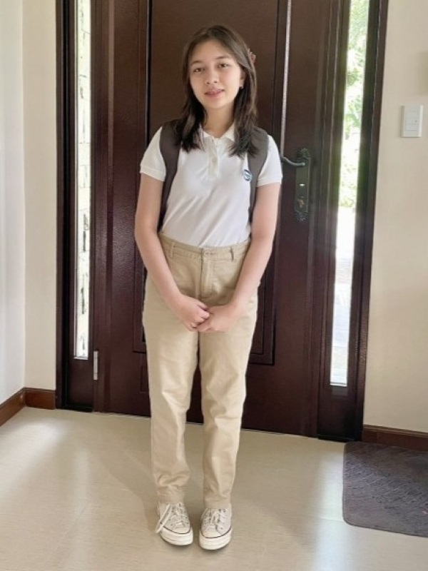 Alba standing inside a doorway on her first day of high school wearing a backpack and school clothes, smiling at the camera; dark wooden door and narrow sidelights behind her, bright natural light and an excited, proud expression