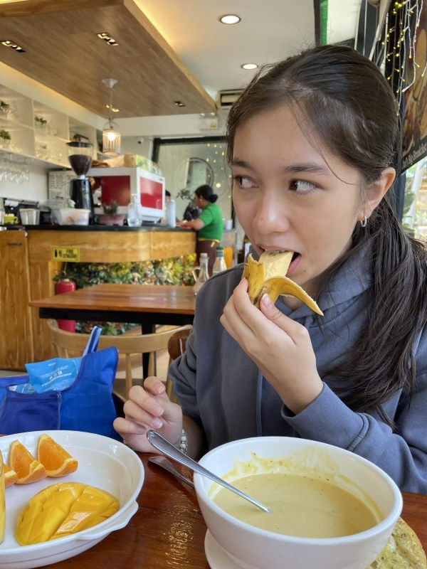 Alba after ballet lesson seated at a cafe table holding a peeled banana to her mouth as she takes a bite; a bowl of creamy soup and a plate with mango and orange slices are on the table; wood counter and staff are visible in the background in a warm, casual cafe setting; she appears relaxed and content; no visible text