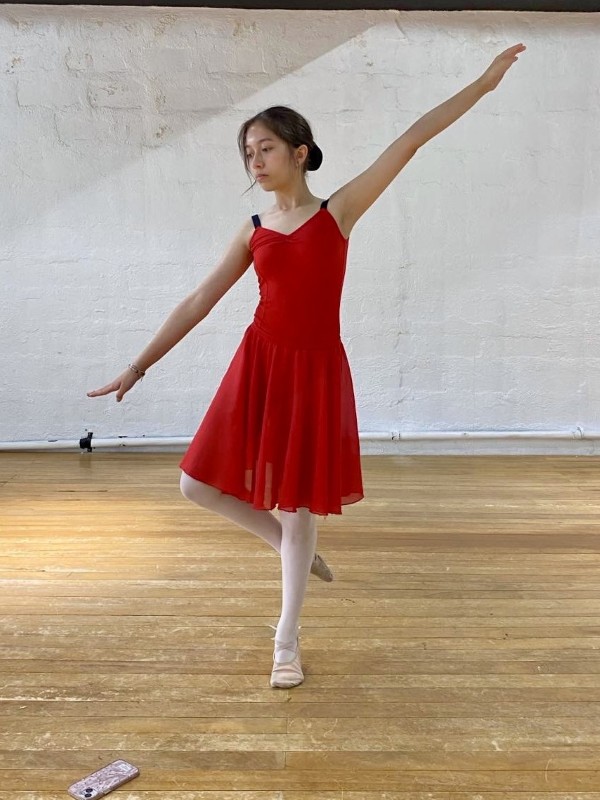 Alba standing on one foot in a graceful pose with arms extended, wearing a red dress; inside a dance studio with a polished wooden floor and white textured wall, soft natural light; mood serene and focused