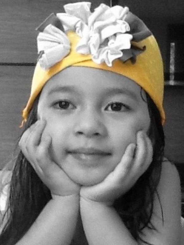 Alba wearing the yellow beanie decorated with layered fabric flowers, resting her chin in her hands and looking at the camera in a close up portrait; black and white photograph with the hat shown in color; indoor setting with a soft, gentle, affectionate mood