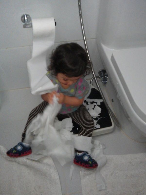 Alba, a toddler sitting on a bathroom floor beside a bathtub and toilet, pulling and tearing toilet paper from a wall-mounted roll with strips strewn across a bath mat and scale; playful and mischievous